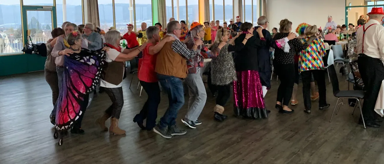 Eine Gruppe von Menschen in verschiedenen Kostümen tanzt gemeinsam in einem großen Raum mit Holzböden und großen Fenstern.