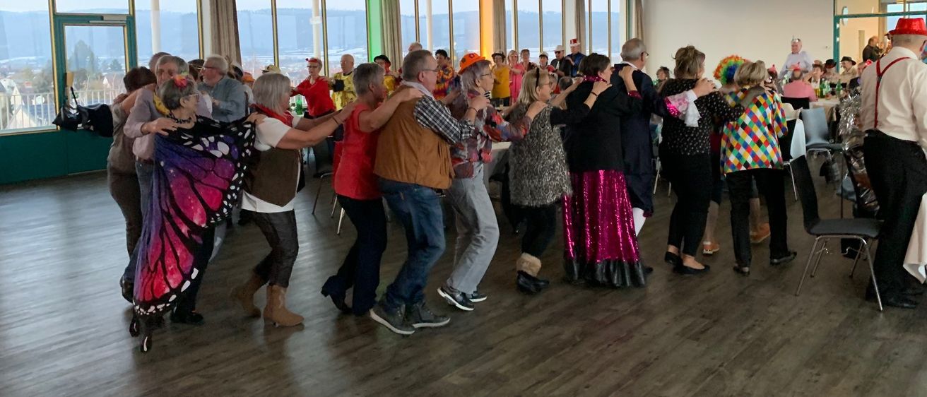 Eine Gruppe von Menschen in verschiedenen Kostümen tanzt gemeinsam in einem großen Raum mit Holzböden und großen Fenstern.