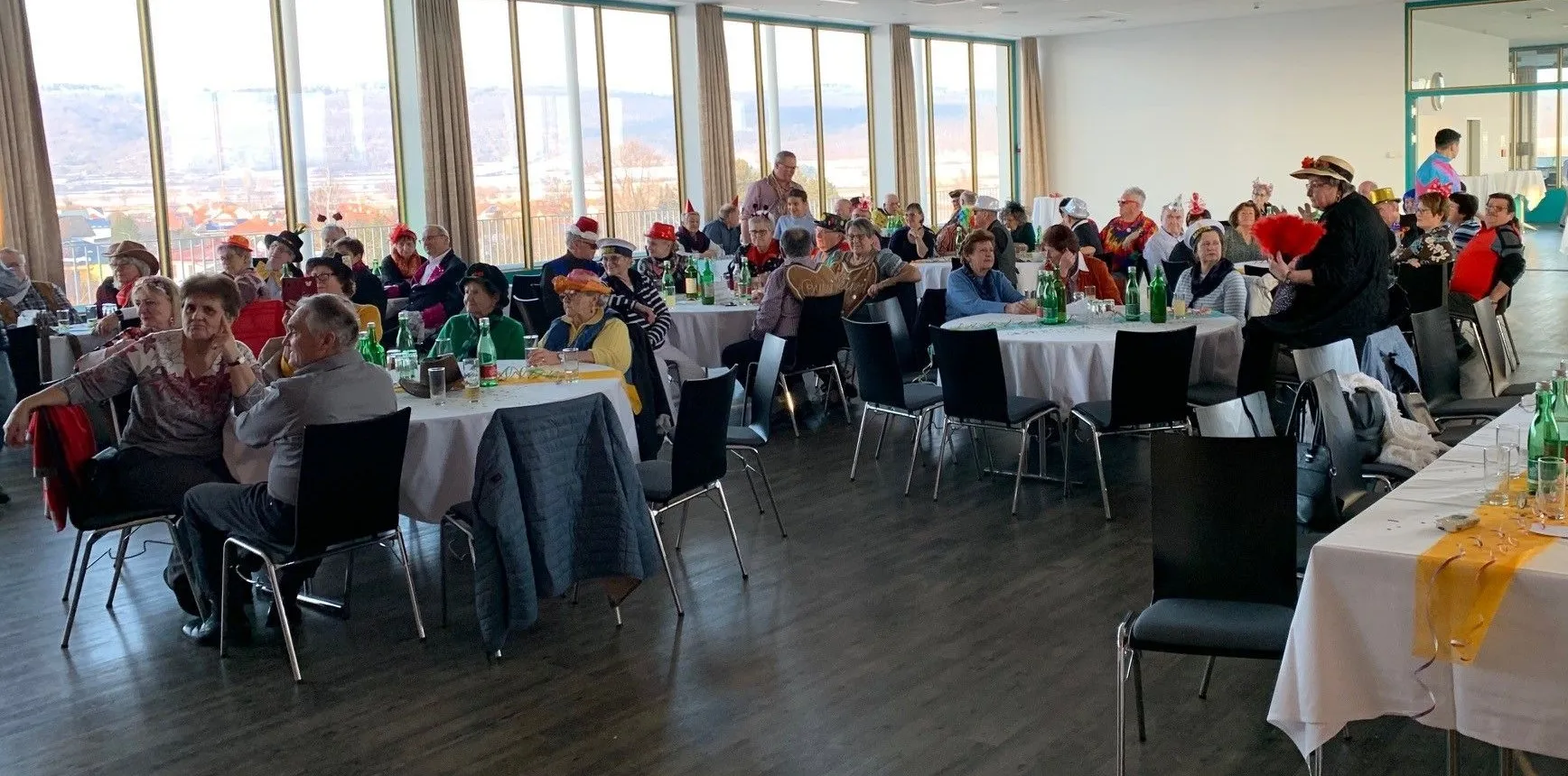Eine Gruppe von Menschen sitzt um Tische in einem Bankettsaal herum und trägt festliche Hüte. Die Tische sind mit Flaschen, Gläsern und anderen Gegenständen geschmückt. Große Fenster bieten einen Blick auf die Landschaft draußen.