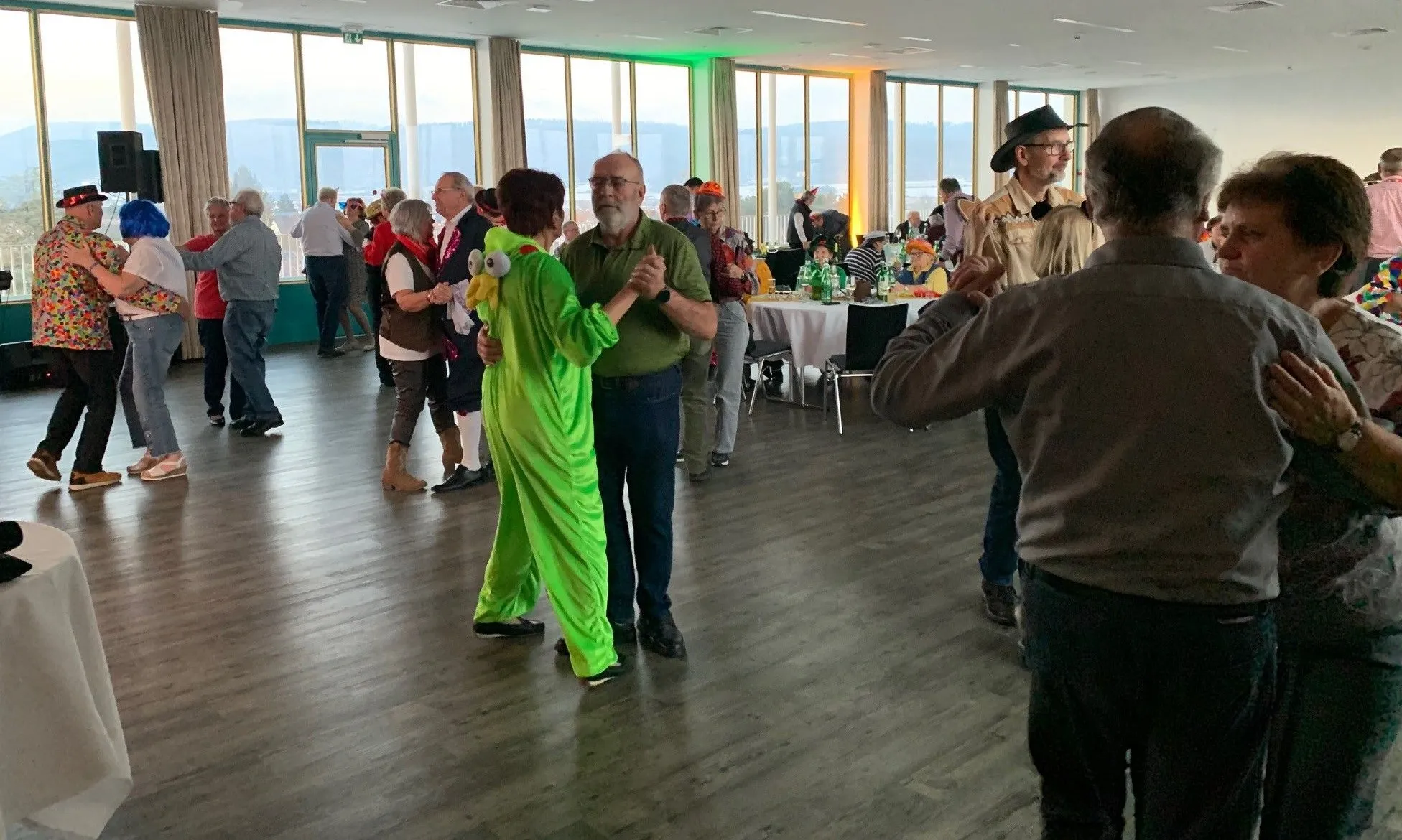 Eine Gruppe von Menschen tanzt in einem Saal mit Holzboden. Einige tragen Kostüme, andere sind lässig gekleidet. Tische mit Essen und Getränken sind aufgestellt. Der Raum hat große Fenster.