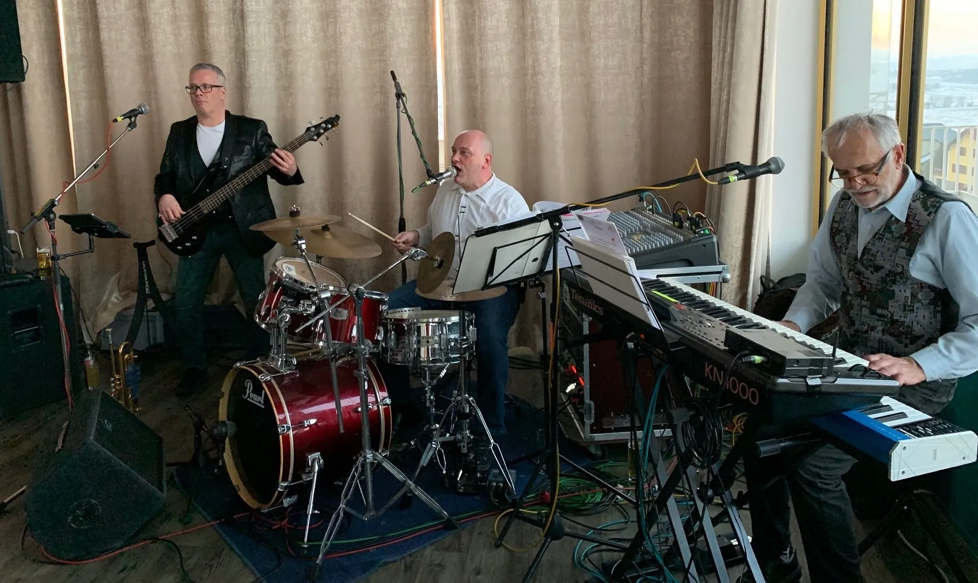 Eine Band spielt, wobei ein Mann die Gitarre, ein anderer das Schlagzeug und eine dritte Person in ein Mikrofon singt.