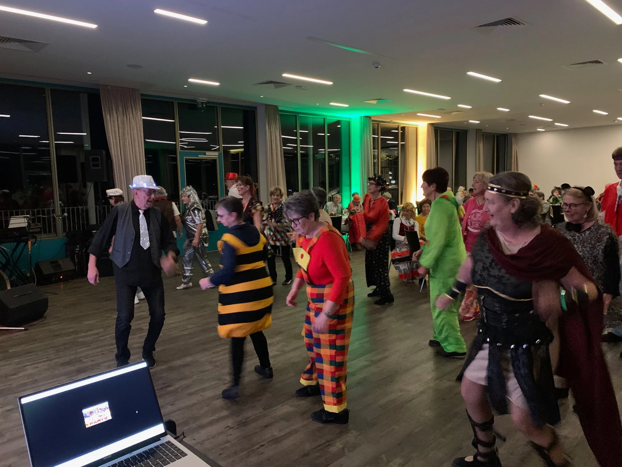 Eine Gruppe von Menschen tanzt in einem Saal, einige in bunten Kostümen. Der Raum hat Holzböden, grüne Lichter und große Fenster. Ein Laptop-Bildschirm ist links sichtbar.
