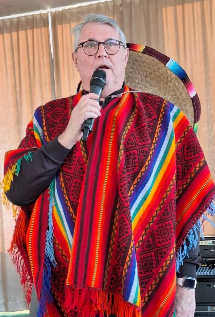 Ein älterer Mann mit Brille spricht in ein Mikrofon, trägt einen farbenfrohen Poncho mit Fransen und einen Hut.