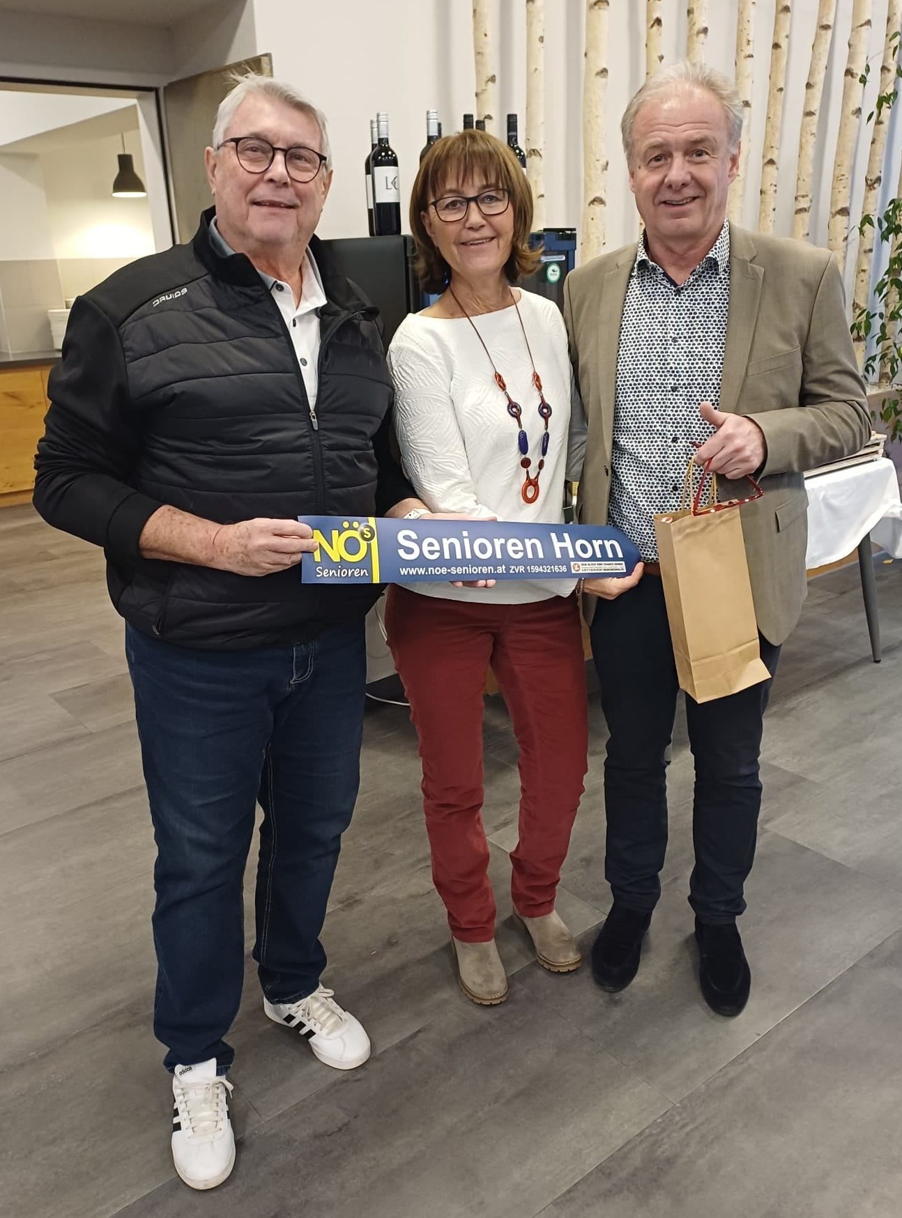 Drei Menschen stehen zusammen und halten ein Schild mit der Aufschrift NO$ Senioren Horn. Die Frau trägt eine Brille und eine Halskette. Die Männer halten Papiertüten und der Mann auf der rechten Seite lächelt.