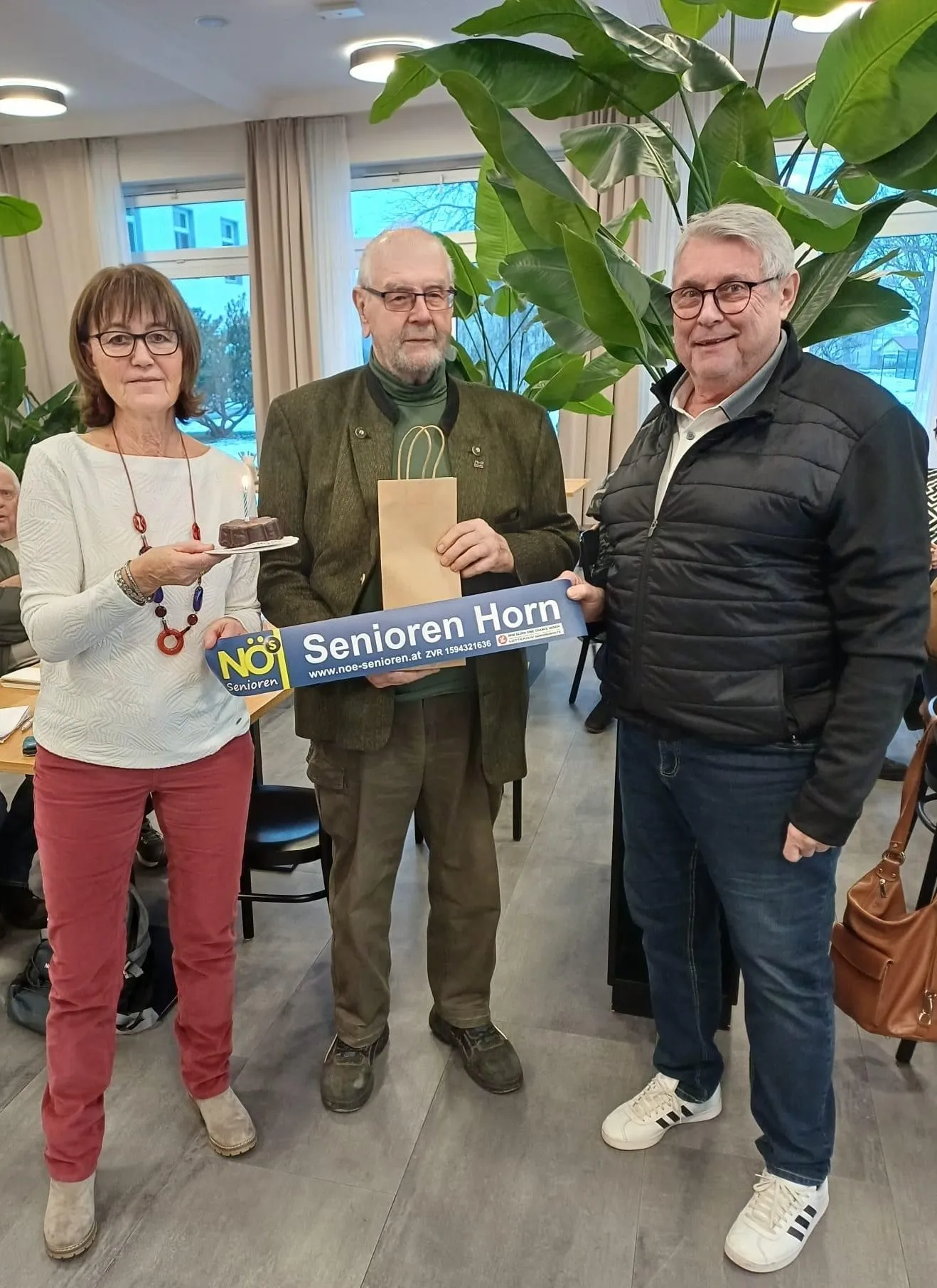 Drei Personen posieren mit einem Kuchen und einem Schild, auf dem 'Senioren Horn' steht. Eine Frau hält den Kuchen, ein Mann das Schild und ein anderer Mann steht neben ihnen. Dahinter befinden sich Pflanzen und ein Tisch.