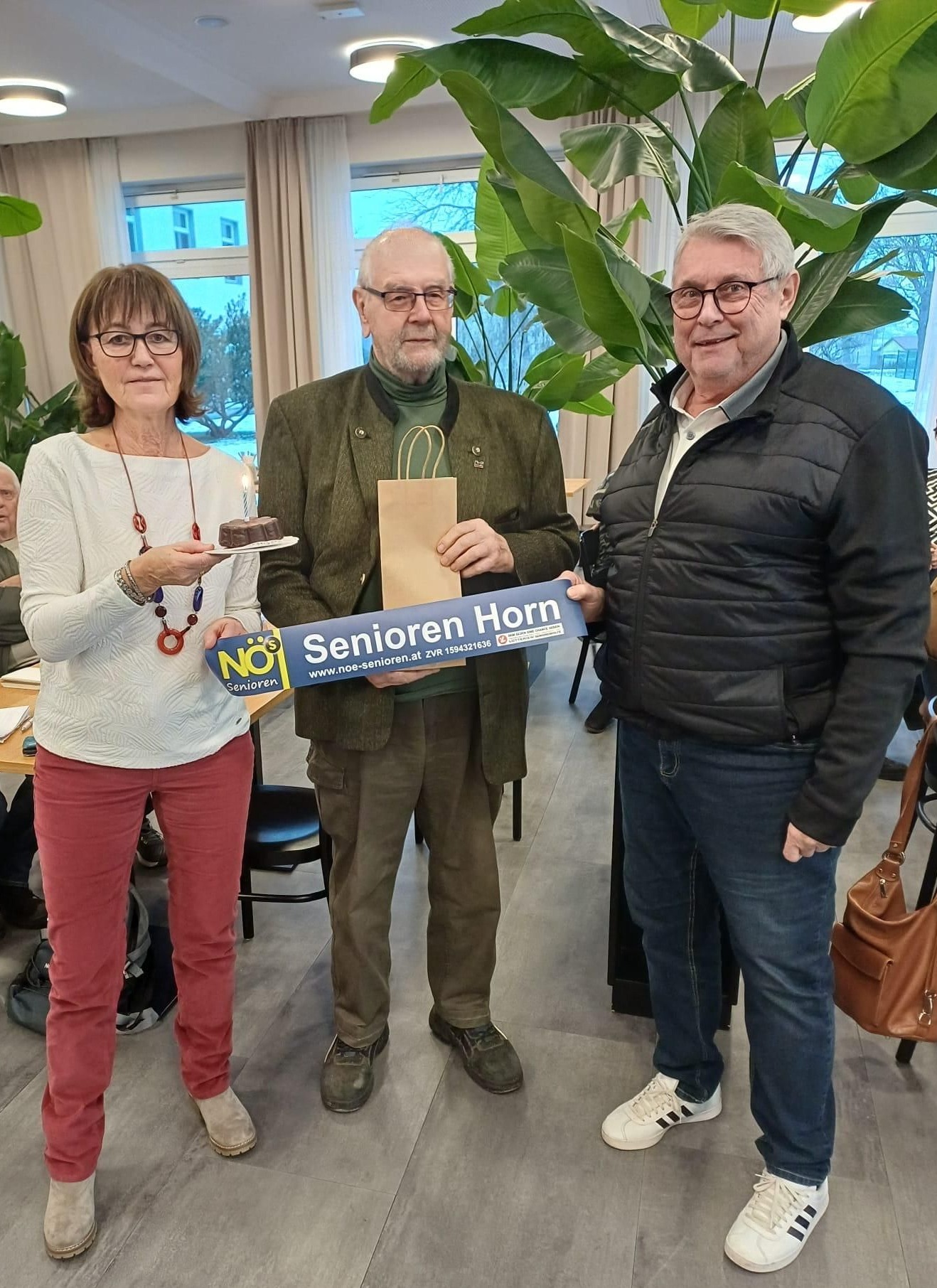 Drei Personen posieren mit einem Kuchen und einem Schild, auf dem 'Senioren Horn' steht. Eine Frau hält den Kuchen, ein Mann das Schild und ein anderer Mann steht neben ihnen. Dahinter befinden sich Pflanzen und ein Tisch.