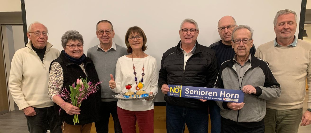 Eine Gruppe älterer Menschen steht zusammen und lächelt für ein Foto. Eine Frau hält Blumen und ein Mann hält ein blaues Schild mit der Aufschrift 'Senioren Horn'.