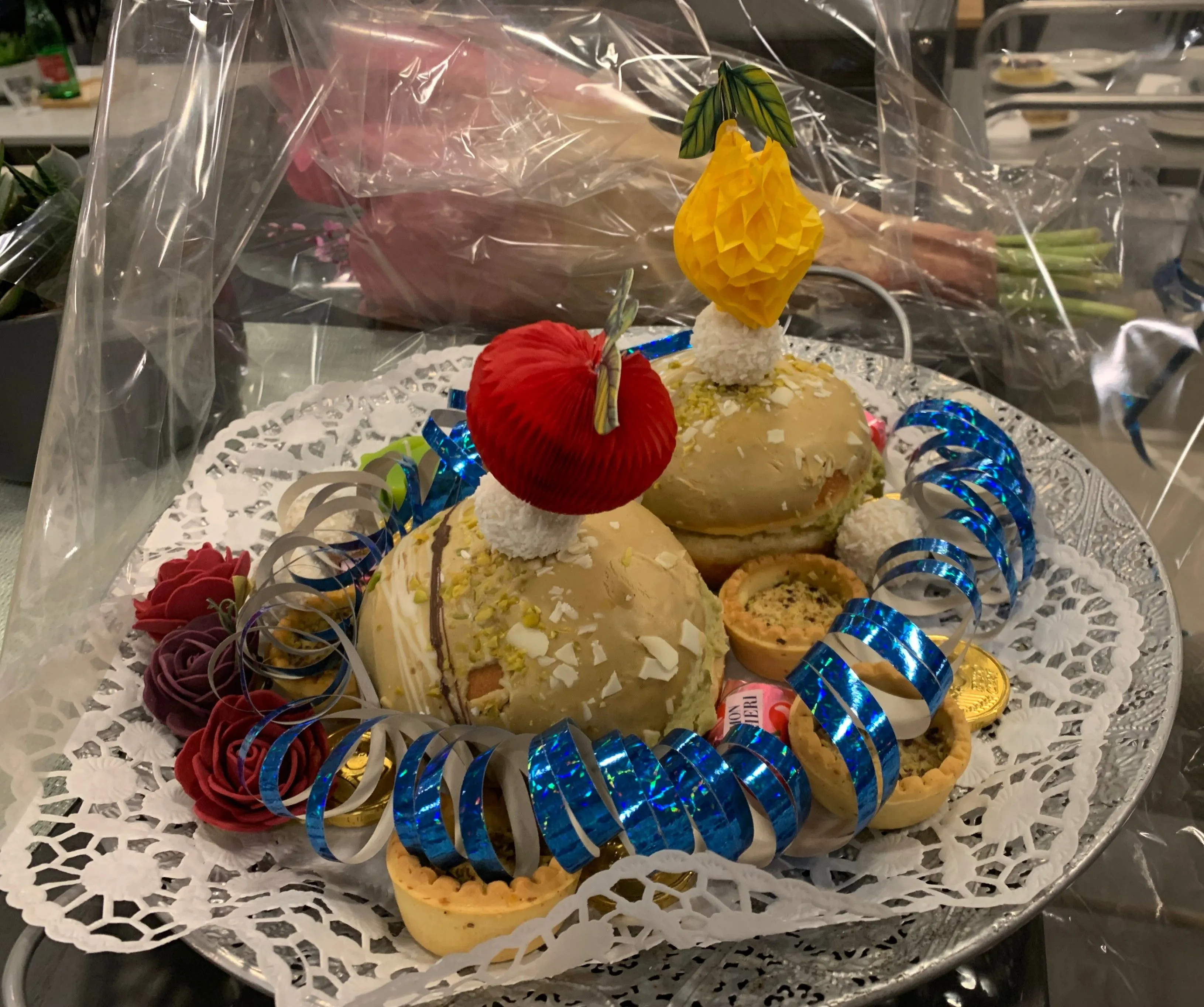 Ein dekorativer Teller zeigt verschiedene Desserts, darunter zwei große Kuchen, die mit einer roten und gelben Frucht gekrönt sind, umgeben von blauen Bändern, Blumen und Miniatur-Törtchen.