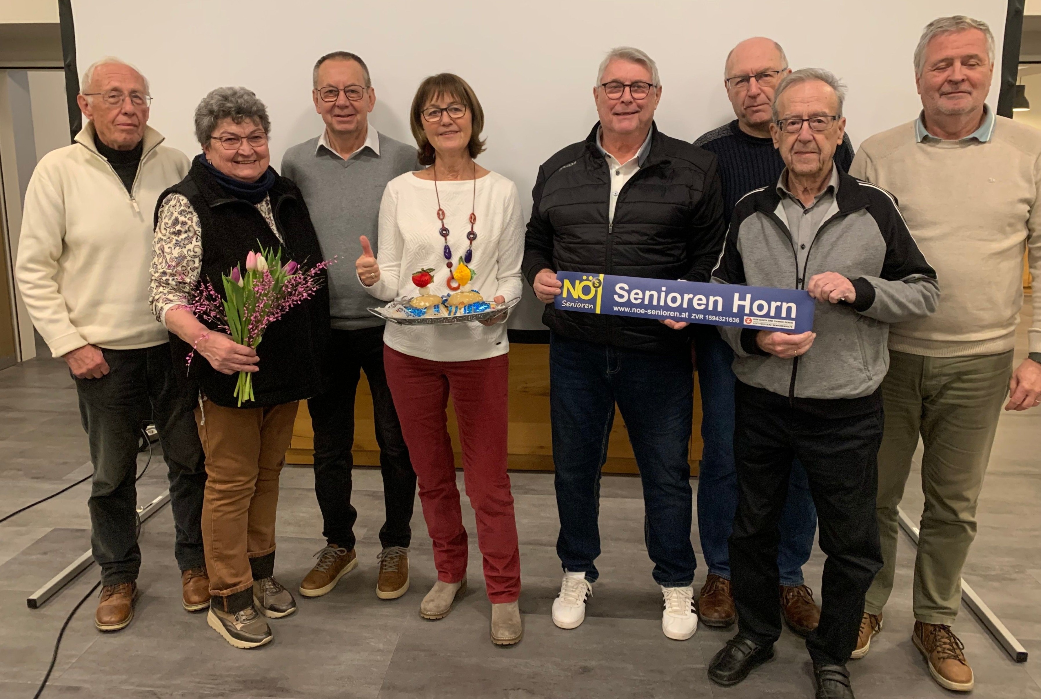 Eine Gruppe älterer Menschen posiert für ein Foto, einer hält ein blaues Schild mit der Aufschrift 'Senioren Horn' und ein anderer hält einen Blumenstrauß. Sie alle lächeln und scheinen ein Ereignis zu feiern.