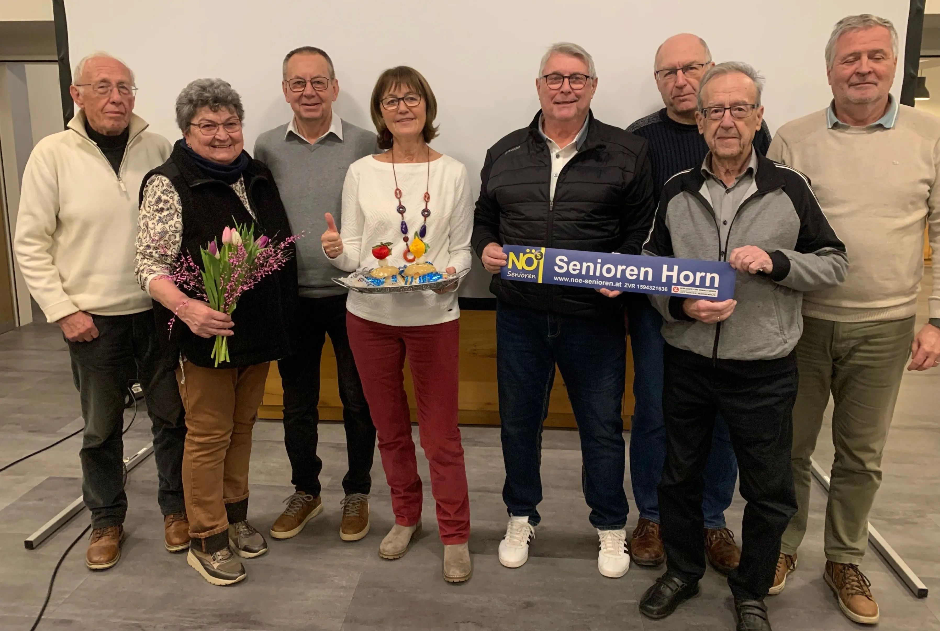 Eine Gruppe älterer Menschen posiert für ein Foto, einer hält ein blaues Schild mit der Aufschrift 'Senioren Horn' und ein anderer hält einen Blumenstrauß. Sie alle lächeln und scheinen ein Ereignis zu feiern.