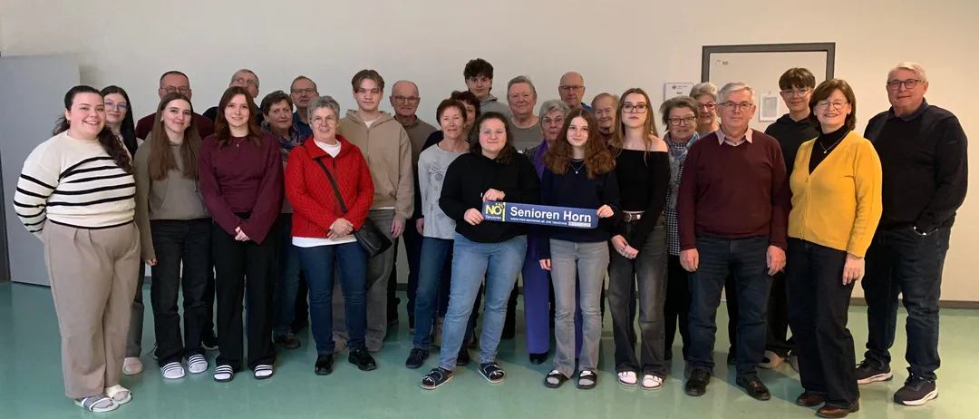 Eine Gruppe von Menschen steht in einem Raum, wobei eine Person ein Banner mit der Aufschrift 'Senioren Horn' hält. Sie sind in verschiedene Kleidungsstücke und Schuhe gekleidet. Der Raum hat eine weiße Wand und einen grünen Boden.