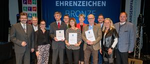 Eine Gruppe von Menschen steht auf einer Bühne, hält Urkunden in den Händen und lächelt für ein Foto. Sie sind von einem blauen Banner mit weißen Text umgeben. Einige tragen Brillen und formelle Kleidung. Die Frau in der Mitte hält eine Urkunde mit ihren Händen.