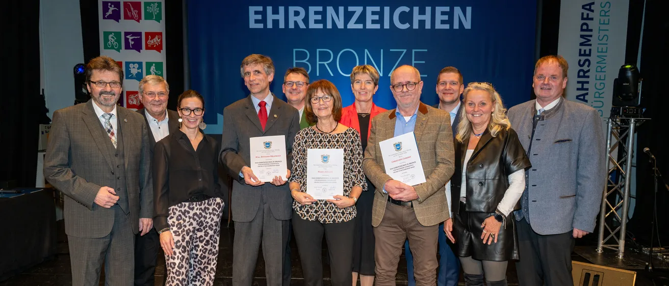 Eine Gruppe von Menschen steht auf einer Bühne, hält Urkunden in den Händen und lächelt für ein Foto. Sie sind von einem blauen Banner mit weißen Text umgeben. Einige tragen Brillen und formelle Kleidung. Die Frau in der Mitte hält eine Urkunde mit ihren Händen.