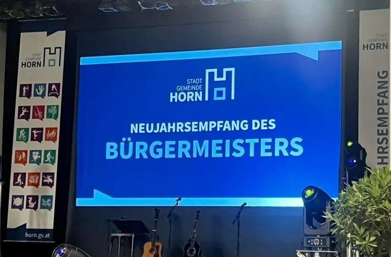 Eine Bühnenaufstellung mit einem großen Bildschirm, auf dem die Worte 'Neujahrsempfang des Bürgermeisters' stehen. Es gibt Banner auf beiden Seiten, Musikinstrumente auf Ständern und eine Scheinwerfer auf der rechten Seite.