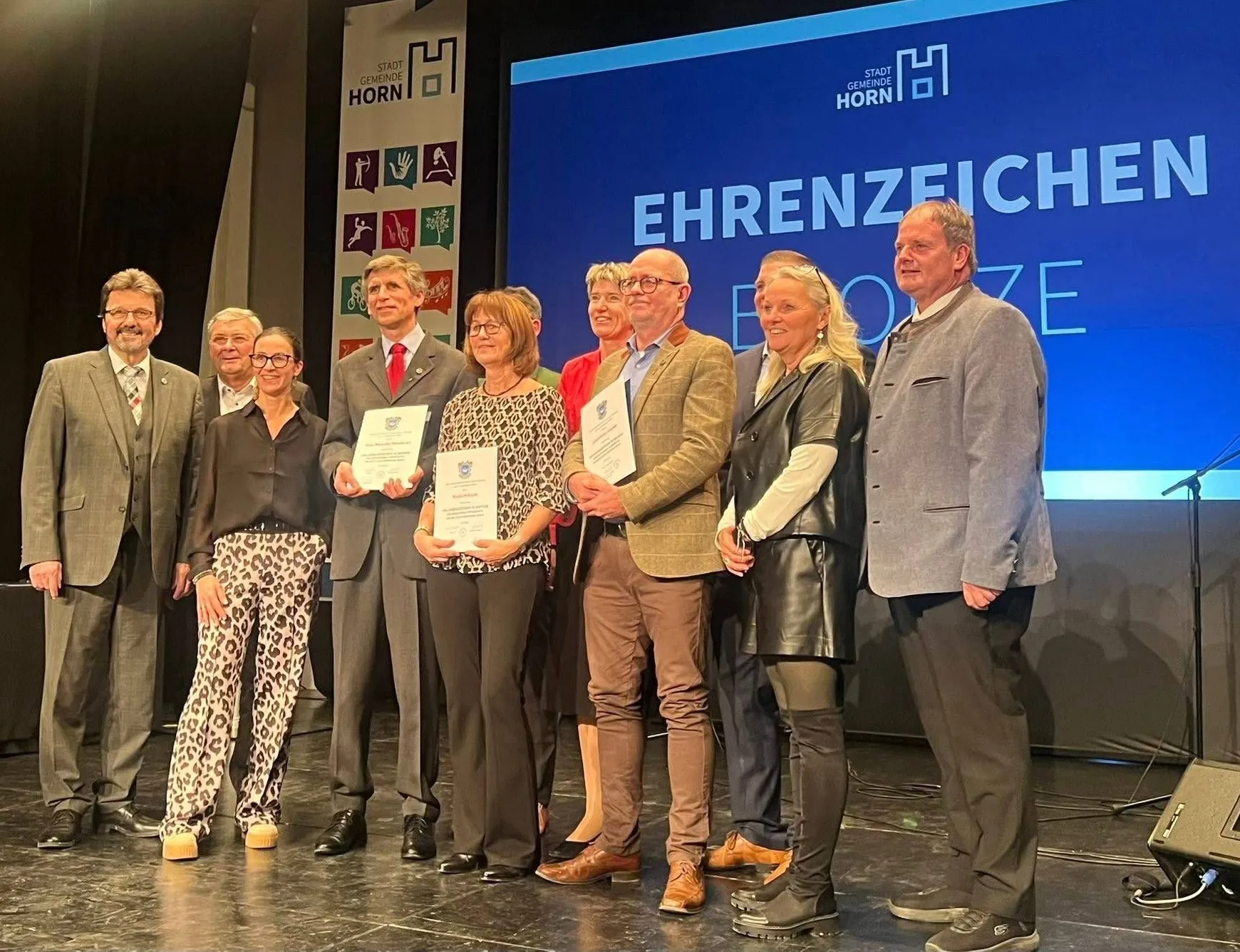 Eine Gruppe von Menschen steht auf einer Bühne und hält Urkunden in den Händen, wobei sich zwei Personen in der Mitte befinden. Hinter ihnen ist eine große blaue Leinwand mit dem Text 'EHRENZEICH'. Sie sind alle formell gekleidet.