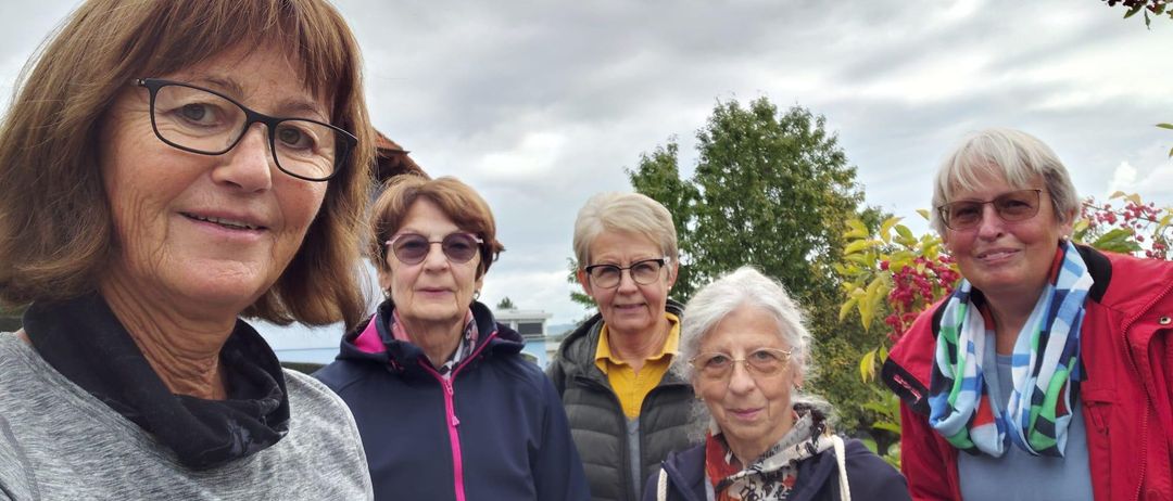 Fünf Frauen stehen im Freien, lächeln und posieren für ein Foto. Sie tragen Jacken und Brillen, eine Frau hält einen Becher. Hinter ihnen steht ein Gebäude und einige Bäume.