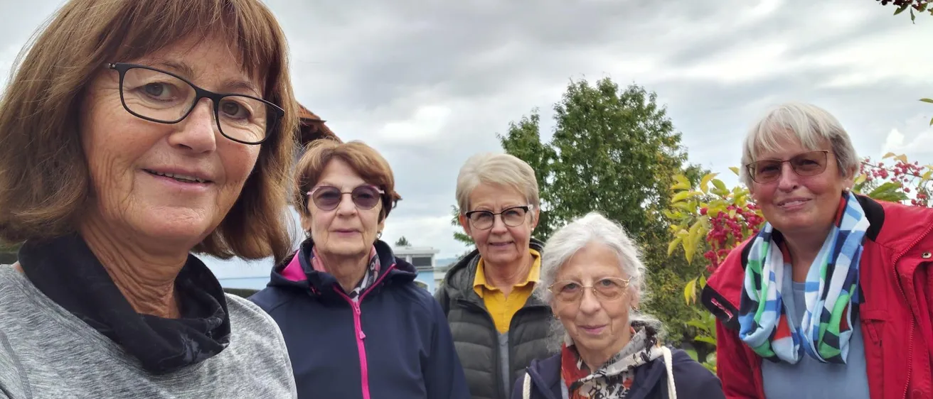 Fünf Frauen stehen im Freien, lächeln und posieren für ein Foto. Sie tragen Jacken und Brillen, eine Frau hält einen Becher. Hinter ihnen steht ein Gebäude und einige Bäume.
