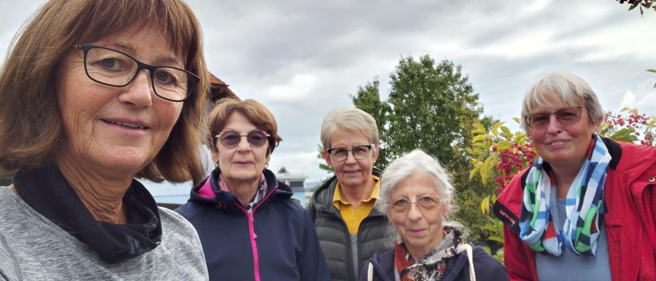 Fünf Frauen stehen im Freien, lächeln und posieren für ein Foto. Sie tragen Jacken und Brillen, eine Frau hält einen Becher. Hinter ihnen steht ein Gebäude und einige Bäume.
