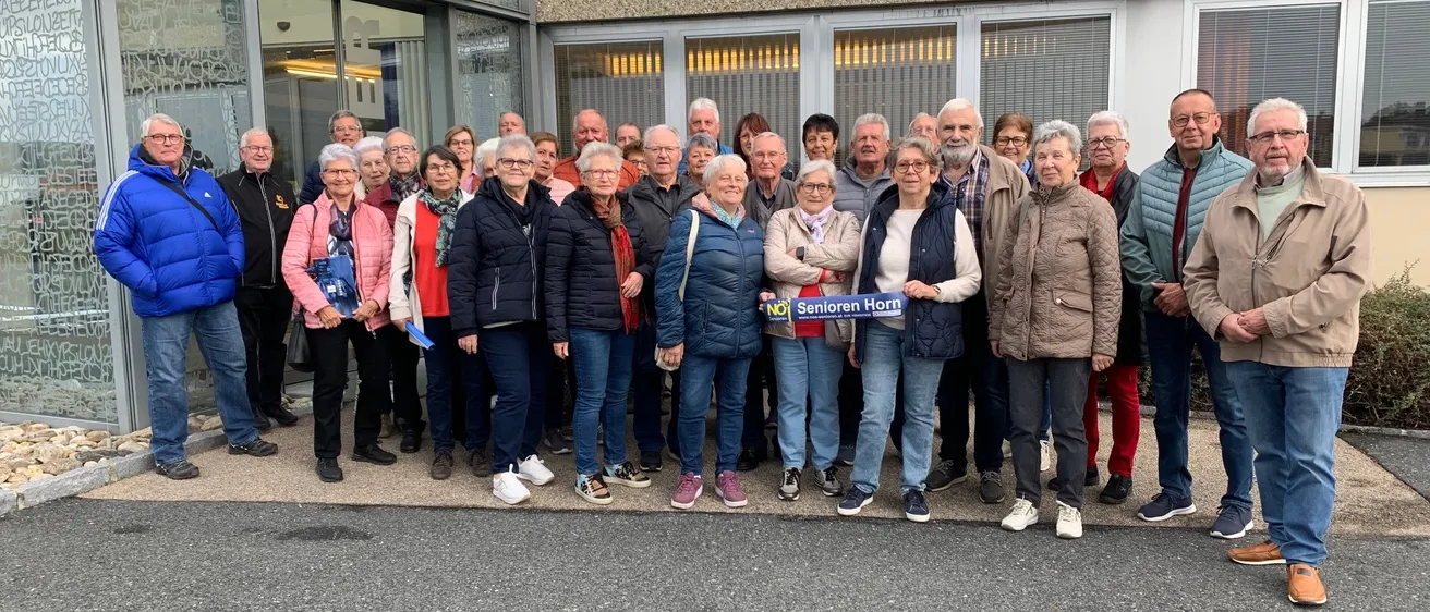 Eine Gruppe älterer Menschen in Jacken und Sneakern steht vor einem Gebäude mit Glasfenstern. Eine Frau hält ein blaues Banner mit den Worten 'Senioren Horn'.