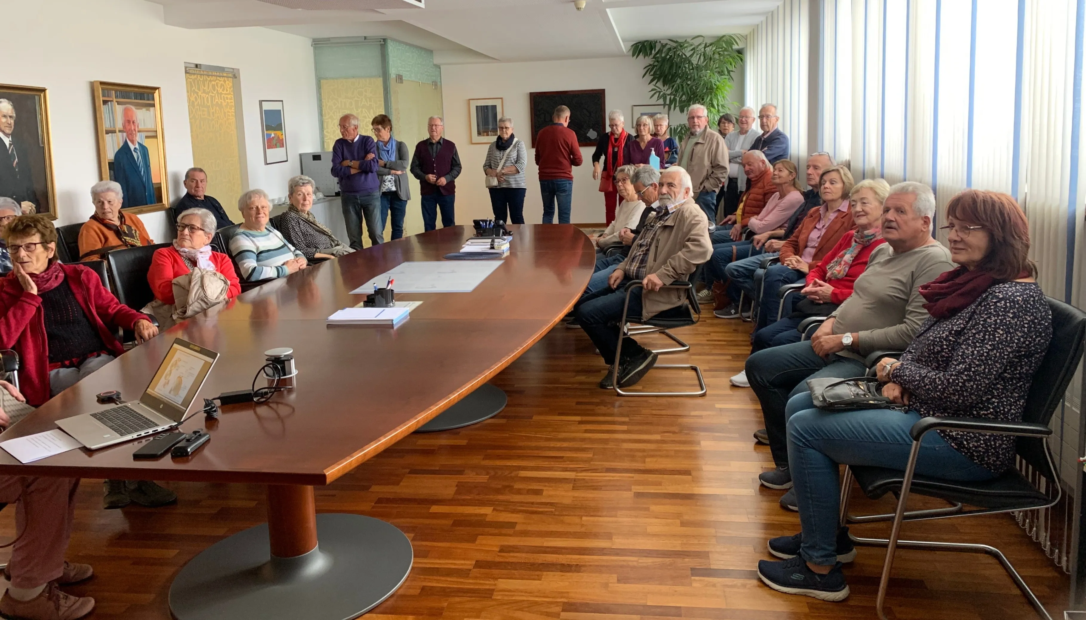 Eine Gruppe von Menschen in einem Besprechungsraum, einige sitzen auf Stühlen und andere stehen in der Nähe eines Tisches mit Papieren und Stiften und blicken nach vorne.