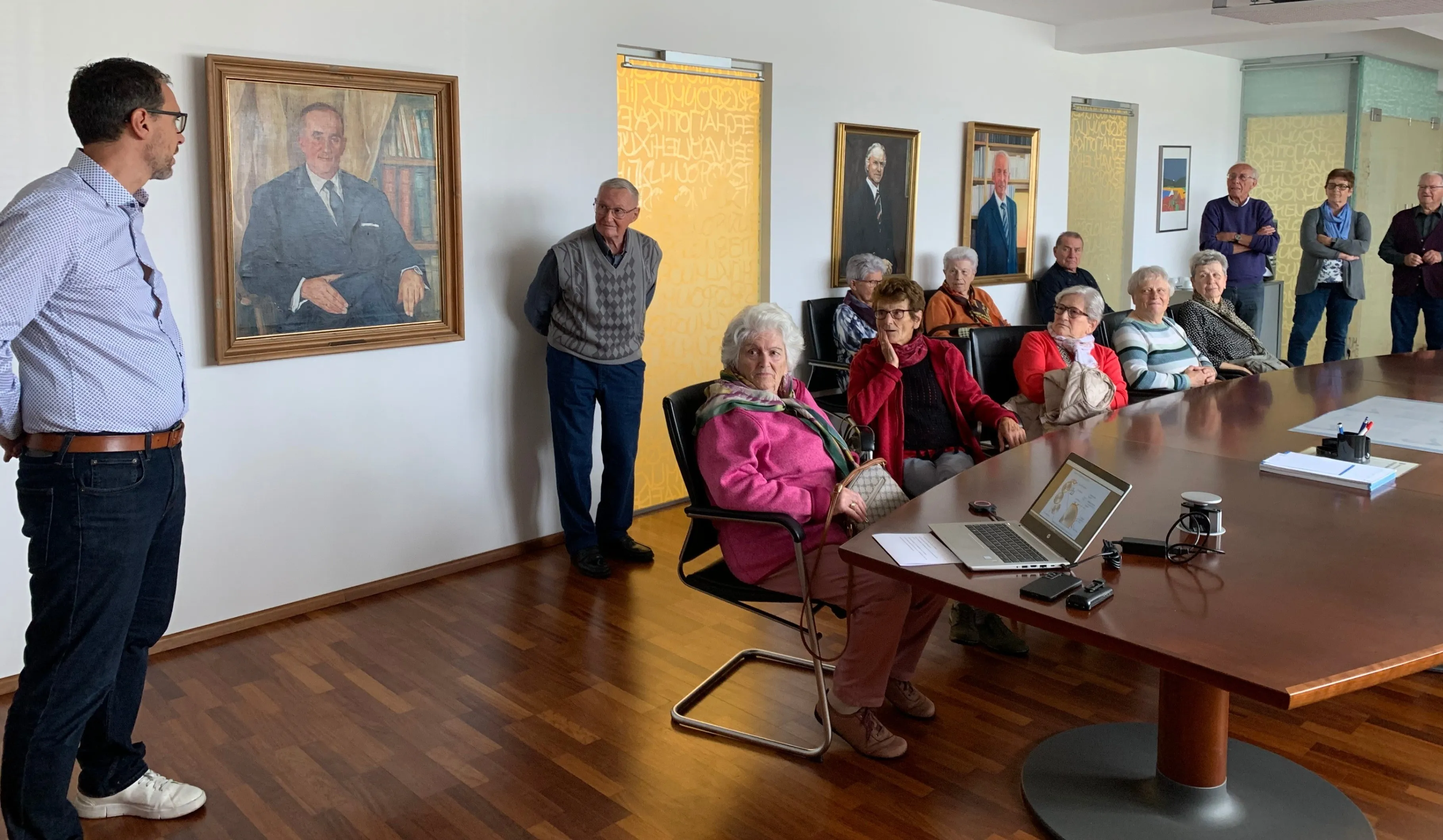 Eine Gruppe älterer Menschen sitzt um einen Tisch mit einem Laptop. Ein älterer Mann steht in der Nähe einer Wand mit einem Porträt.