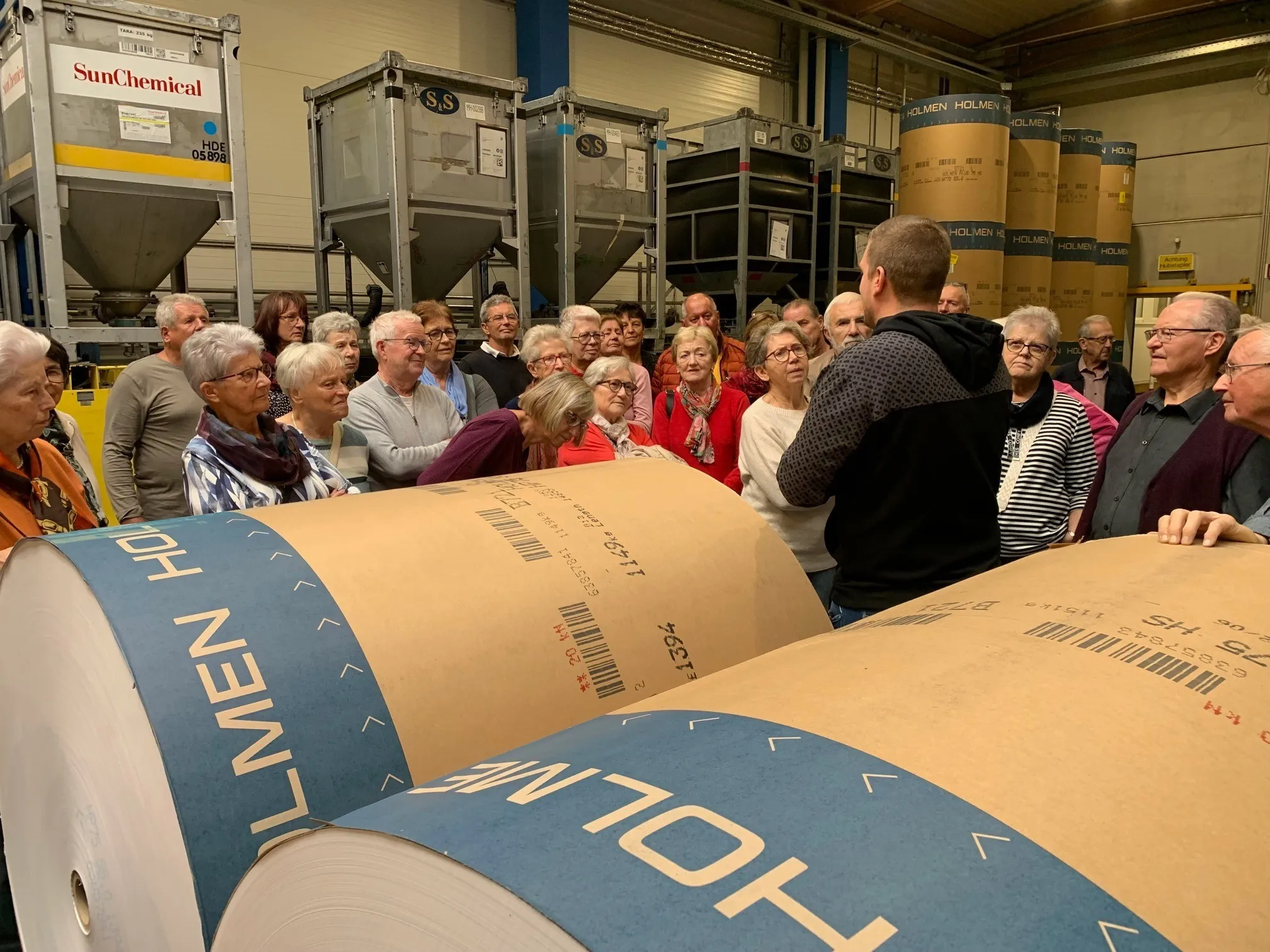 Eine Gruppe von Menschen ist um eine große Papierrolle in einer Fabrik versammelt und hört einem Mann zu, der spricht. Dahinter sind weitere Rollen und Metallstrukturen zu sehen.
