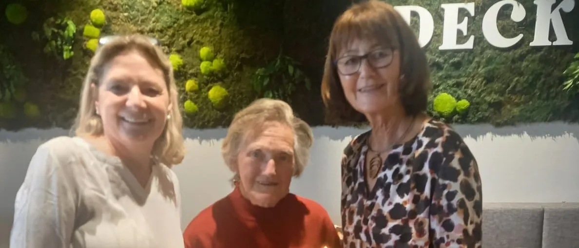 Drei Frauen stehen zusammen, eine hält einen Kuchen, die Frau in der Mitte bläst eine Kerze aus. Hinter ihnen ist eine Wand mit grünem Moos und Text.