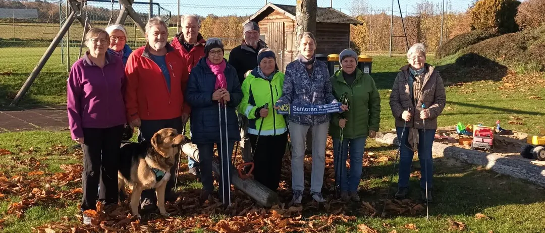 Eine Gruppe von Menschen und ein Hund posiert für ein Foto in einem Park mit Bäumen, Gras und einem Spielplatz im Hintergrund.