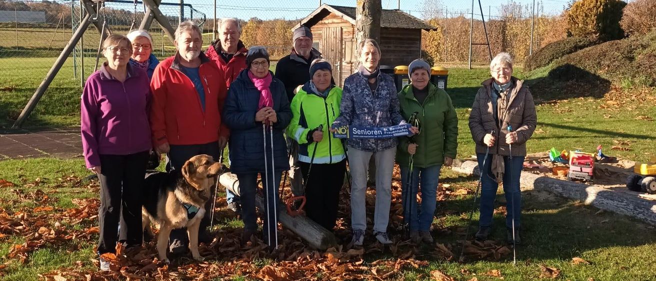Eine Gruppe von Menschen und ein Hund posiert für ein Foto in einem Park mit Bäumen, Gras und einem Spielplatz im Hintergrund.