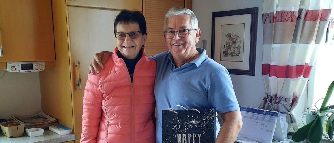 Ein Mann und eine Frau stehen lächelnd in einer Küche, mit einer Geburtstagskarte. Hinter ihnen befinden sich Schränke und ein gerahmtes Bild an der Wand.