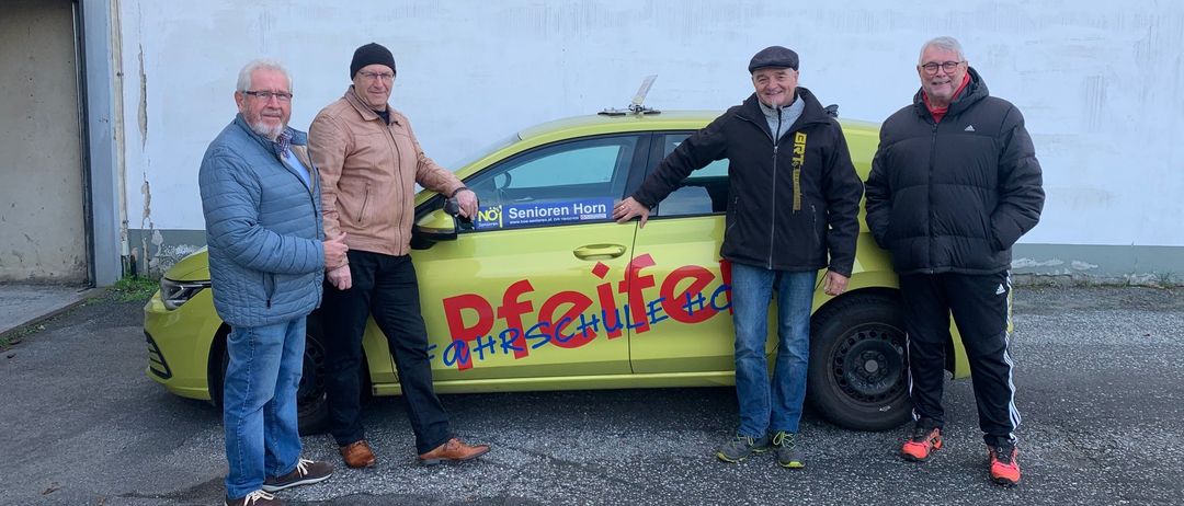 Vier Männer stehen vor einem gelben Auto, auf dem 'Pfeifenschule' steht. Sie tragen alle Winterkleidung und lächeln für die Kamera.