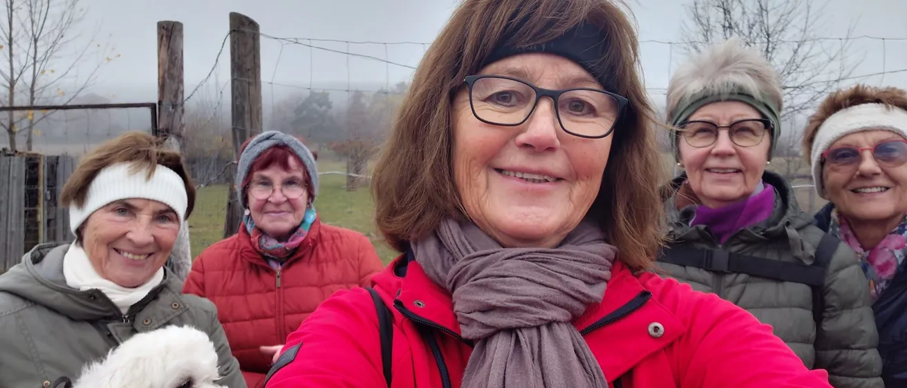 Fünf Frauen lächeln in einem nebligen Außenbereich. Die Frau vorne trägt einen roten Mantel und eine Schürze. Ein weißer Hund ist links. Dahinter ist ein Zaun und Bäume.