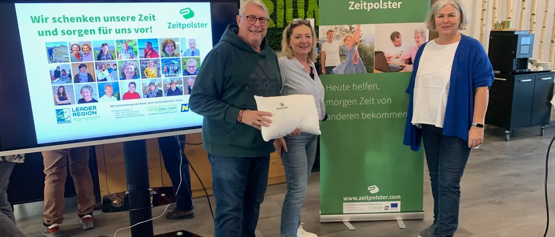 Drei Menschen stehen vor einem großen Bildschirm mit einem Zeitpolster-Banner und halten ein Kissen. Ein lächelnder älterer Mann trägt eine grüne Kapuzenjacke, eine Frau mit Brille ein weißes Hemd und eine andere Frau eine blaue Strickjacke. Hinter ihnen zeigt ein großer Bildschirm verschiedene Fotos.