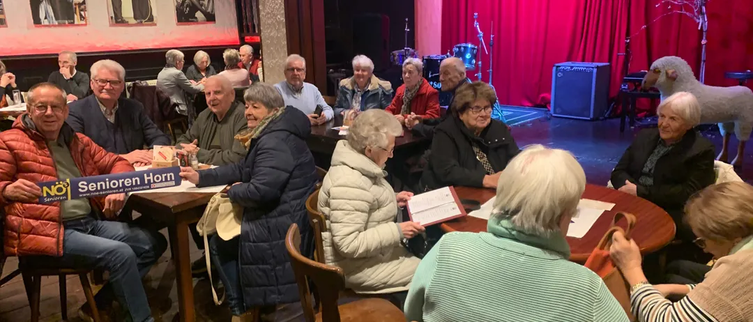 Eine Gruppe älterer Menschen sitzt in einem Raum mit Holzböden an Tischen. Einige schauen auf Papiere. Ein roter Vorhang ist im Hintergrund.