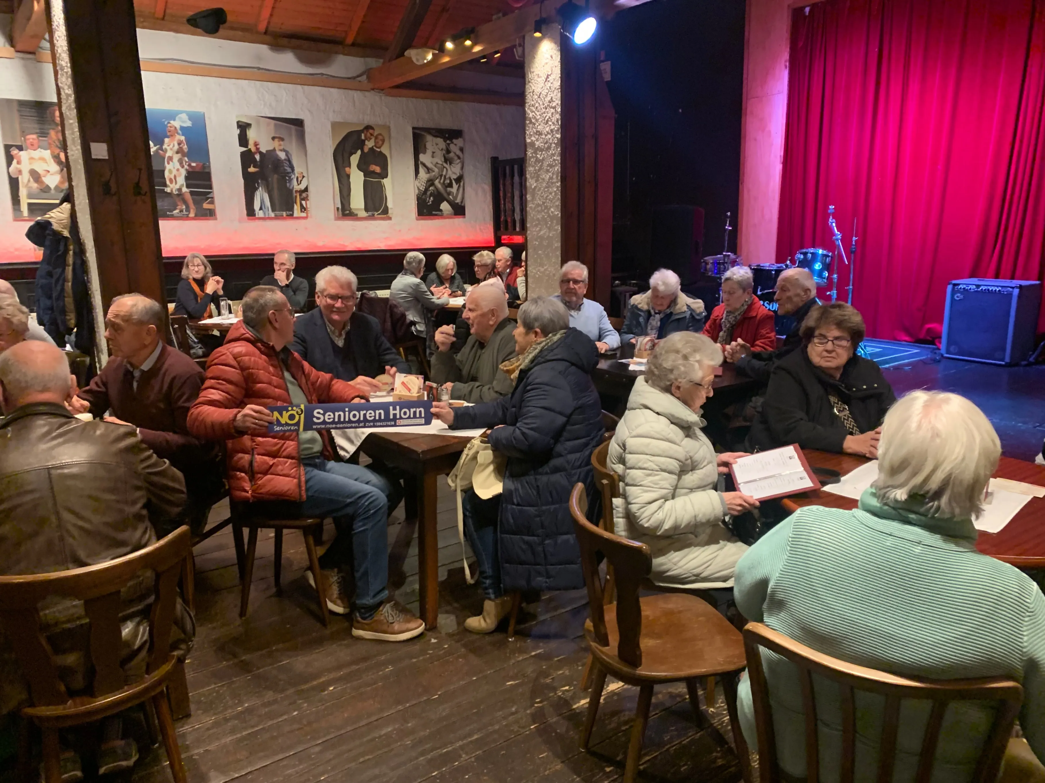 In einem Restaurant sitzen mehrere ältere Menschen an Tischen, einige lesen. Ein Schild mit 'Senioren Horn' ist sichtbar. Der Raum hat einen Holzboden, Stühle und dekorative Bilder an der Wand.