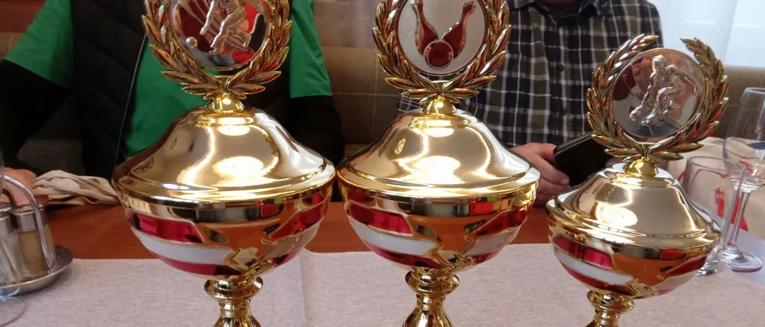 Drei goldene Pokale mit rot-weißer Verzierung stehen auf einem Tisch. Jeder Pokal hat eine metallische Krone und eine Plakette mit Platzierungen. Ein Mann in einem karierten Hemd ist im Hintergrund zu sehen.
