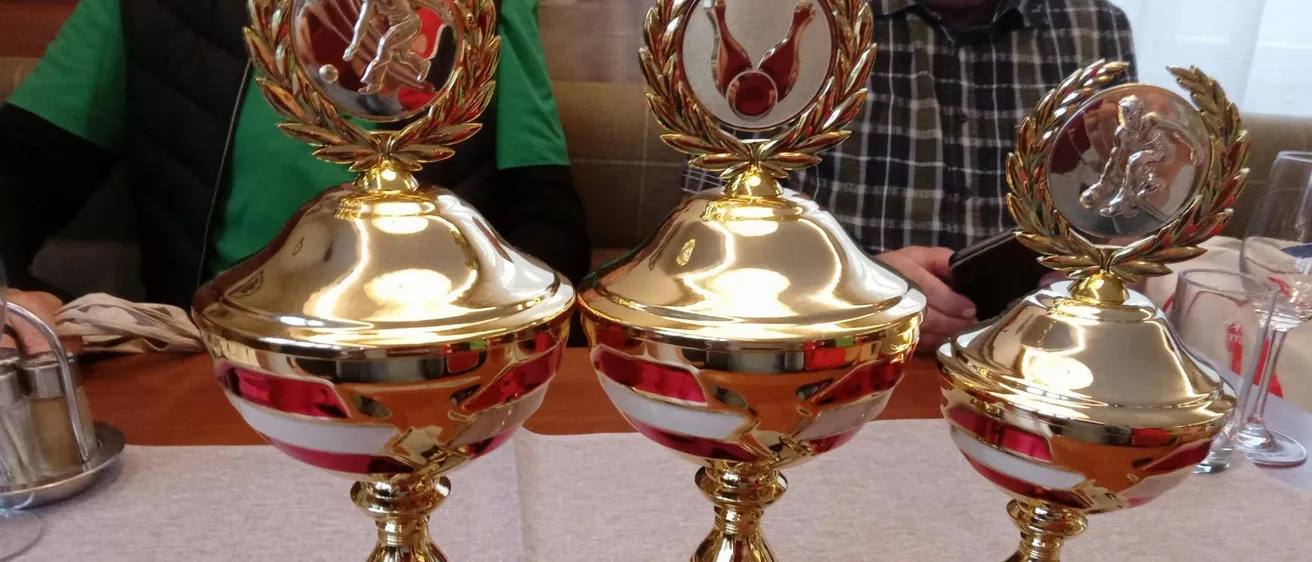 Drei goldene Pokale mit rot-weißer Verzierung stehen auf einem Tisch. Jeder Pokal hat eine metallische Krone und eine Plakette mit Platzierungen. Ein Mann in einem karierten Hemd ist im Hintergrund zu sehen.