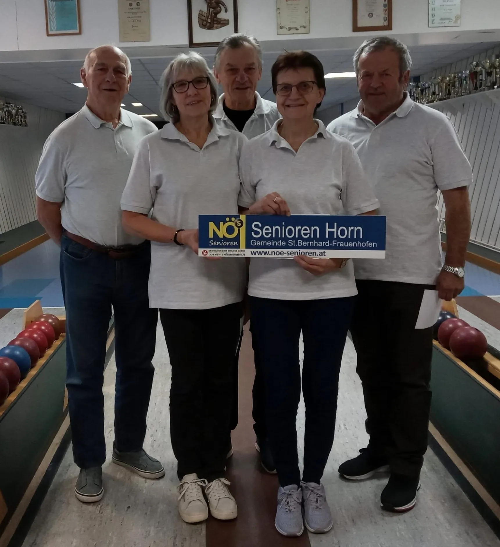 Fünf Personen in passenden Shirts posieren mit einem blauen Schild, halten eine Plakette und lächeln für ein Foto in einer Bowlingbahn.