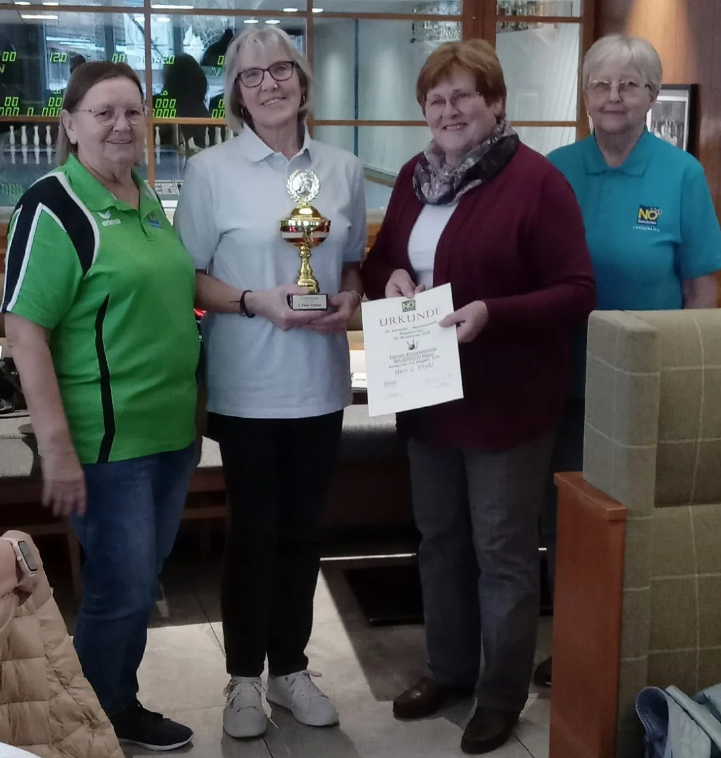 Vier Frauen stehen zusammen und halten einen Pokal und ein Zertifikat. Die Frau in der Mitte hält ein Zertifikat mit 'URKUNDE' darauf. Dahinter befindet sich eine Bowlingbahn.