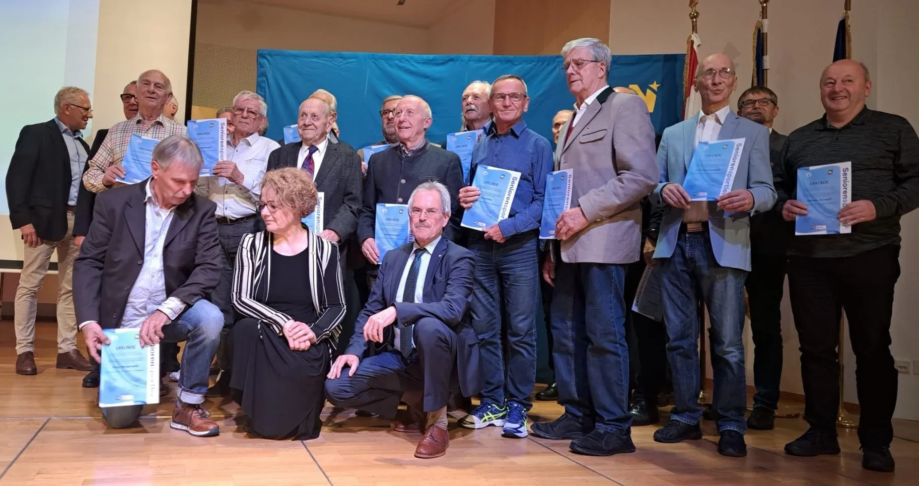 Eine Gruppe älterer Männer und Frauen posiert für ein Foto mit Urkunden. Sie sind formell gekleidet, einige in Anzügen und Krawatten, andere in Freizeitkleidung. Der Hintergrund ist blau.