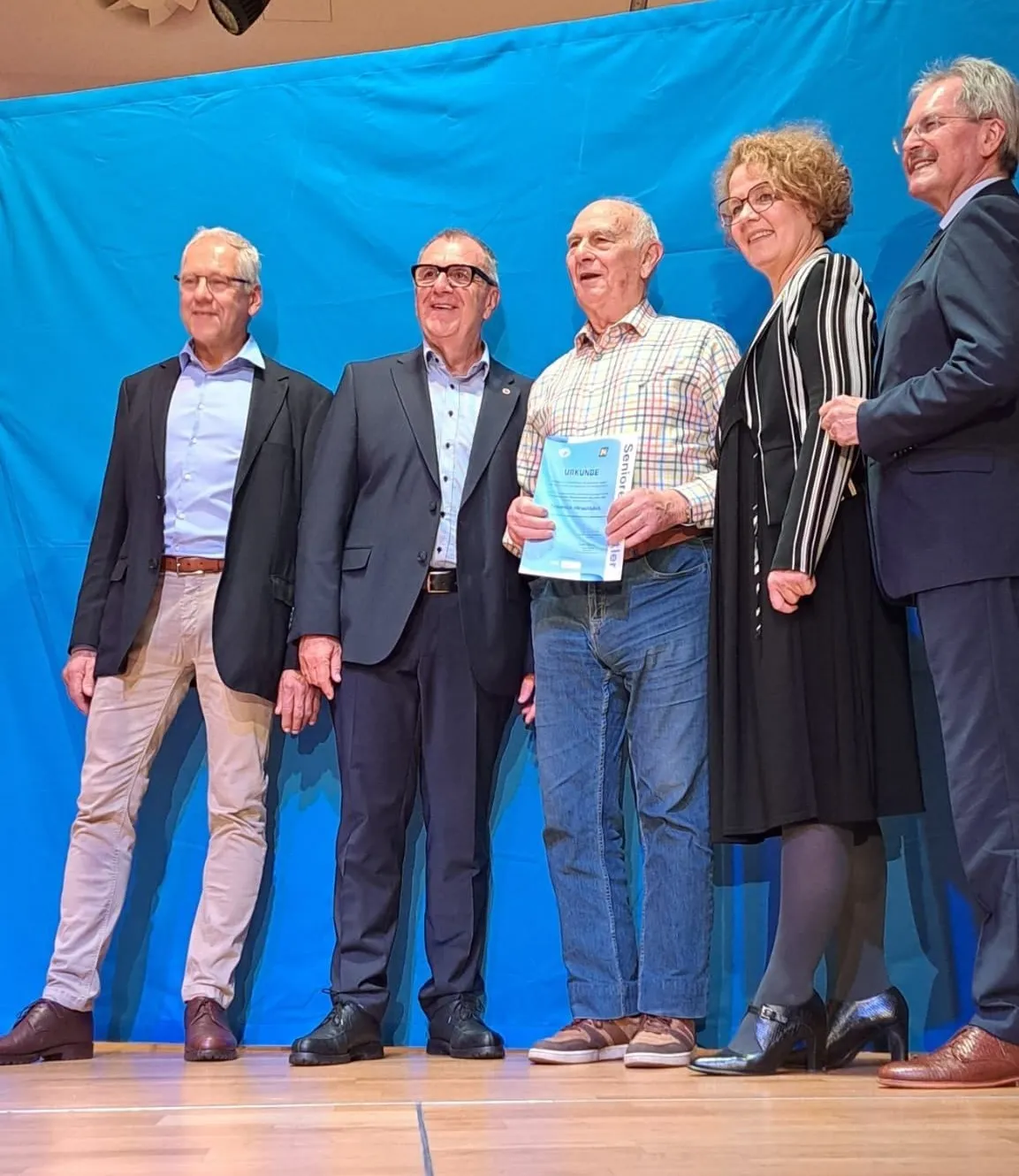Fünf Personen stehen auf einer Bühne vor einem blauen Hintergrund. Ein Mann hält ein Zertifikat und lächelt. Ein Mann neben ihm trägt eine Brille. Eine Frau neben ihm hat eine Brille und einen gestreiften Mantel. Zwei andere Männer stehen an den Seiten.