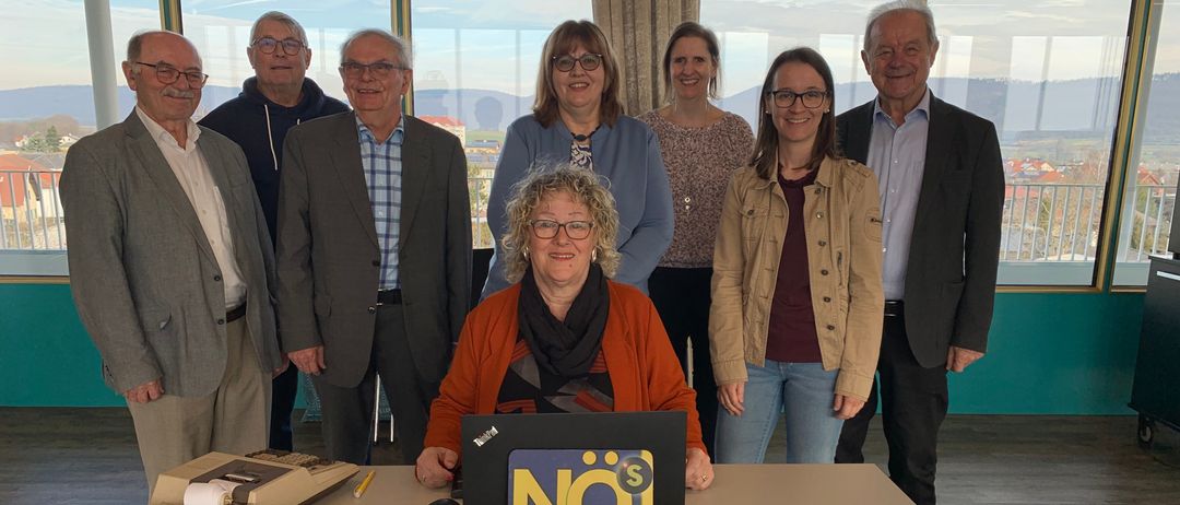 Eine Gruppe von Menschen steht hinter einem Tisch mit einem Laptop, auf dem die Wörter 'NO$ Senioren' stehen. Papiere liegen auf dem Tisch und eine Taschenrechnerin befindet sich auf der linken Seite. Sie befinden sich in einem Raum mit großen Fenstern.