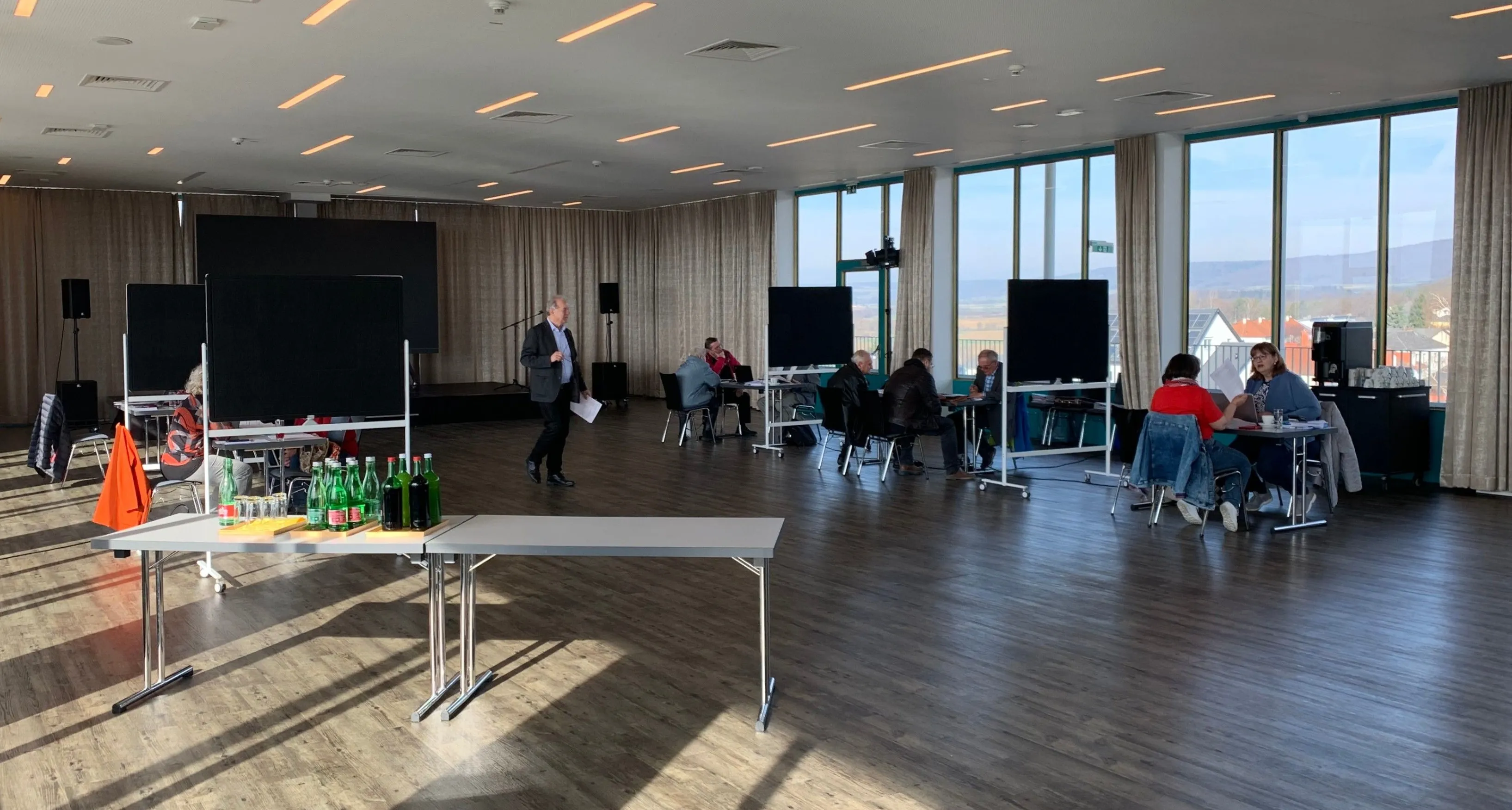 Ein Konferenzraum mit großen Fenstern, einer Bühne und Menschen, die an Tischen sitzen. Ein Mann steht neben einem Tisch mit Flaschen.