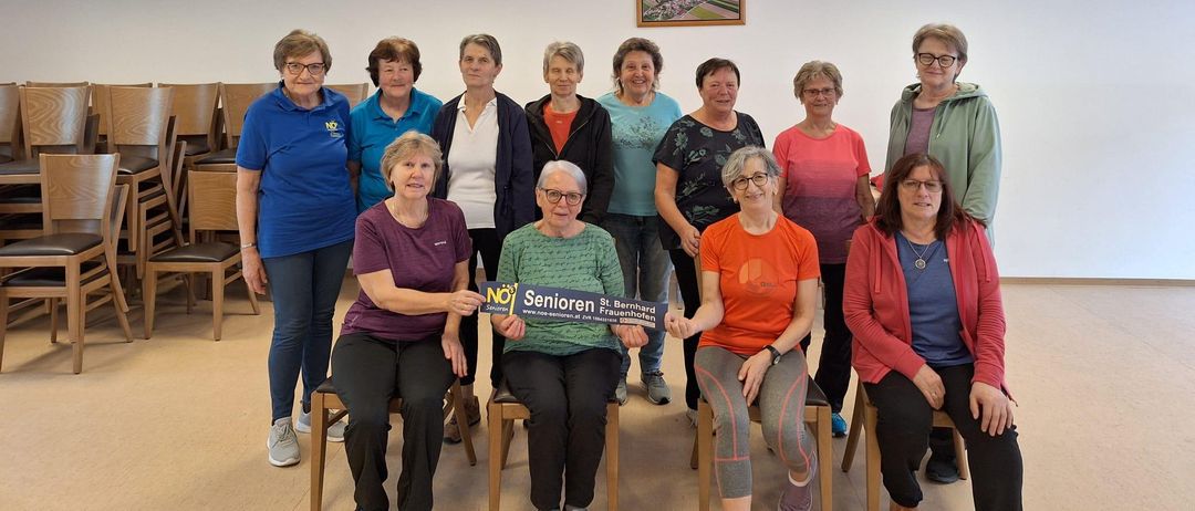 Eine Gruppe von Frauen posiert für ein Foto in einem Raum. Eine Frau hält ein blaues Schild mit der Aufschrift 'Senioren St. Bernhard Frauenhofen.' Die Frauen tragen Freizeitkleidung und sitzen auf Stühlen. Der Raum hat weiße Wände und einen Bilderrahmen an der Wand.