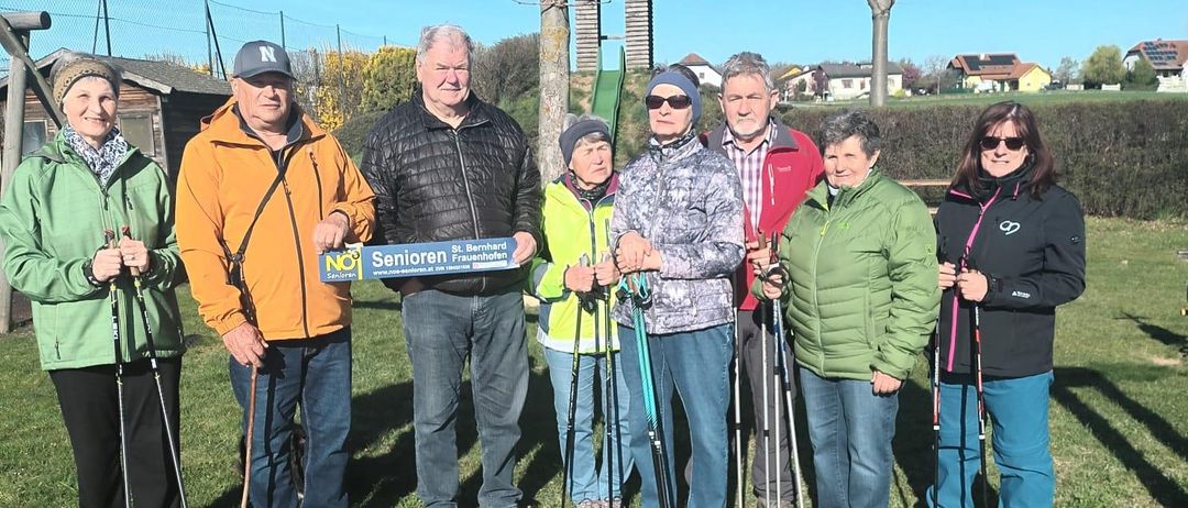 Sieben Menschen stehen auf dem Rasen mit Wanderstöcken. Sie lächeln und scheinen für ein Foto zu posieren. Eine Fahne ist in der Mitte.
