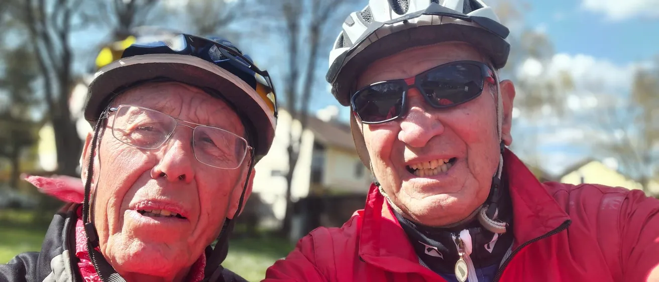Zwei ältere Männer, beide mit Fahrradhelmen, lächeln und posieren für ein Selfie. Der Mann auf der linken Seite hat einen Schal um den Hals. Dahinter sind Bäume und ein Haus zu sehen.