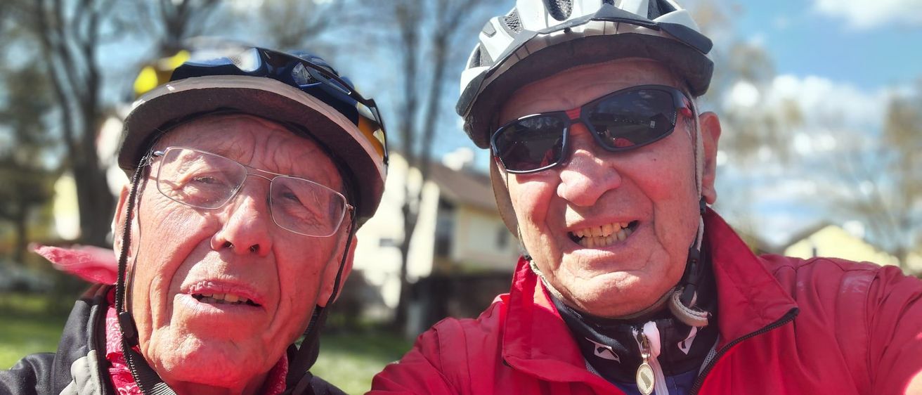 Zwei ältere Männer, beide mit Fahrradhelmen, lächeln und posieren für ein Selfie. Der Mann auf der linken Seite hat einen Schal um den Hals. Dahinter sind Bäume und ein Haus zu sehen.
