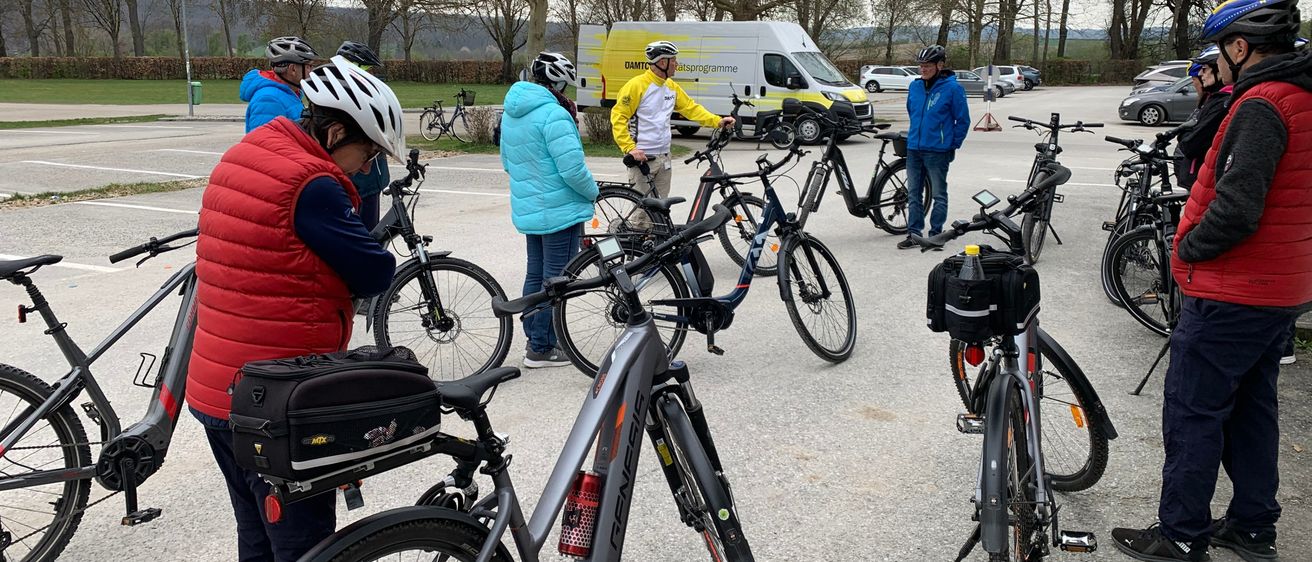 Eine Gruppe von Radfahrern versammelt sich auf einem Parkplatz und bereitet ihre Fahrräder vor. Ein Mann in einem gelben Jackett steht in der Nähe, und ein Van ist hinter ihnen geparkt. Bäume und eine entfernte Bergkette sind sichtbar.