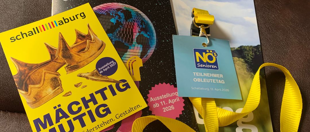 Ein gelbes Buch mit dem Titel Machtig Mutig, Broschüren und ein gelber Lanyard mit einem Schild, das NOs Teilnehmer Obleutetag auf einer braunen Oberfläche liest.