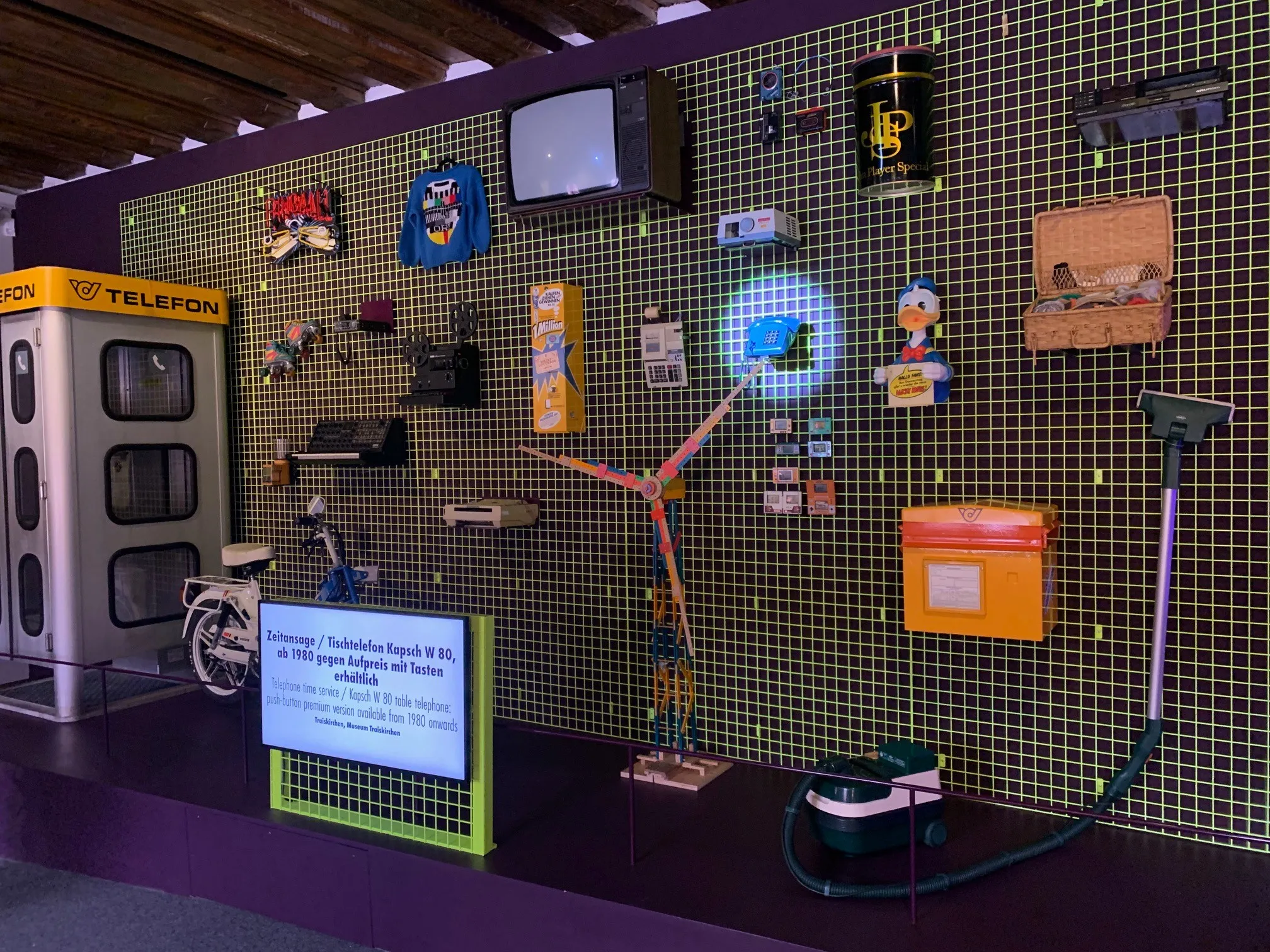 Eine Ausstellung mit einer Mischung aus alten und modernen Gegenständen. Ein blaues Schild mit Informationen über eine Ausstellung, ein Fahrrad, ein Staubsauger und verschiedene Objekte wie ein Fernseher, ein Spielzeug und eine Schachtel.